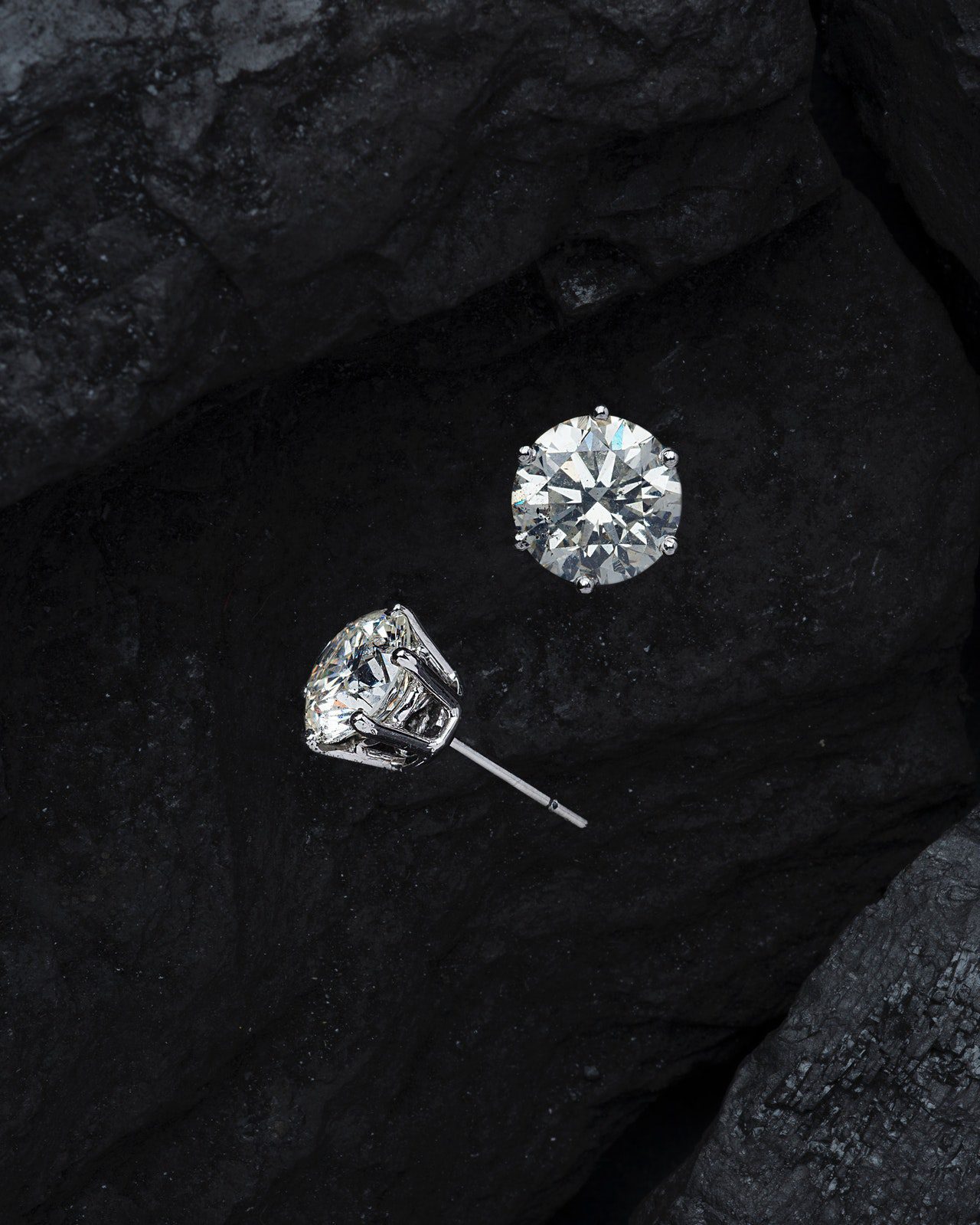 Two round diamond stud earrings, one upright and one on its side, resting on dark, textured rocks.