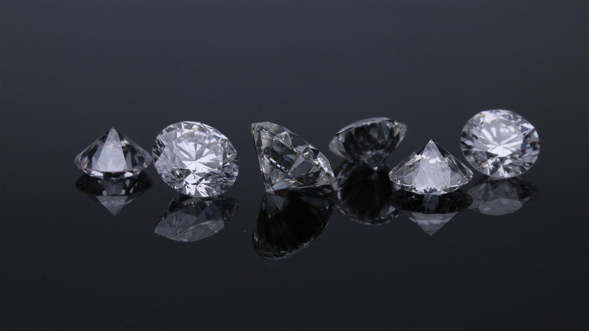 Seven brilliant-cut diamonds with reflections on a dark, reflective surface.