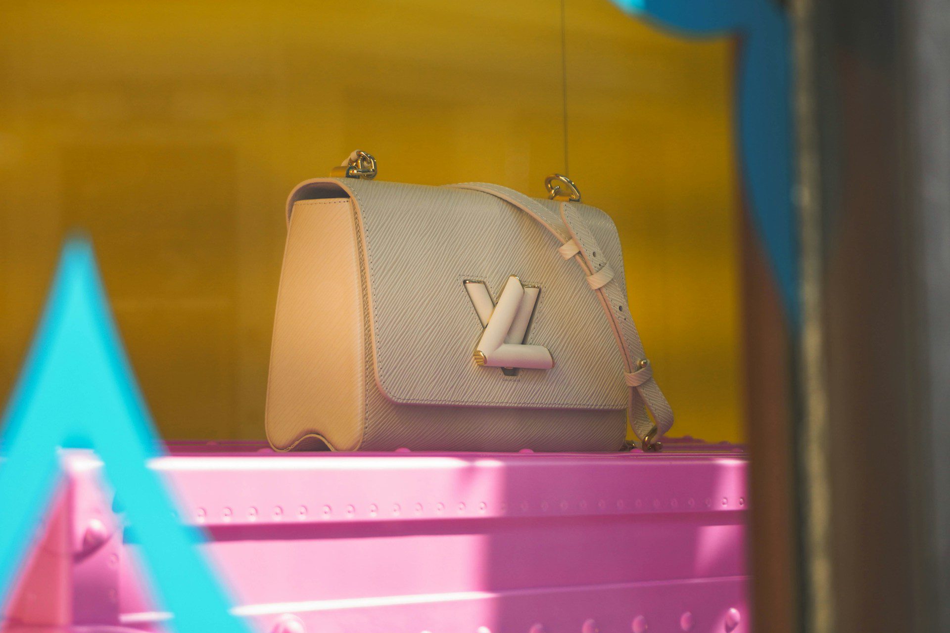 A light pink designer handbag with a prominent silver logo sits on a vibrant pink display shelf.