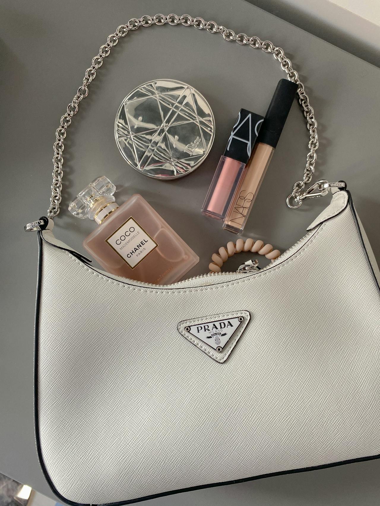 A white Prada shoulder bag with a silver chain strap, perfume, makeup, and a bracelet on a gray surface.