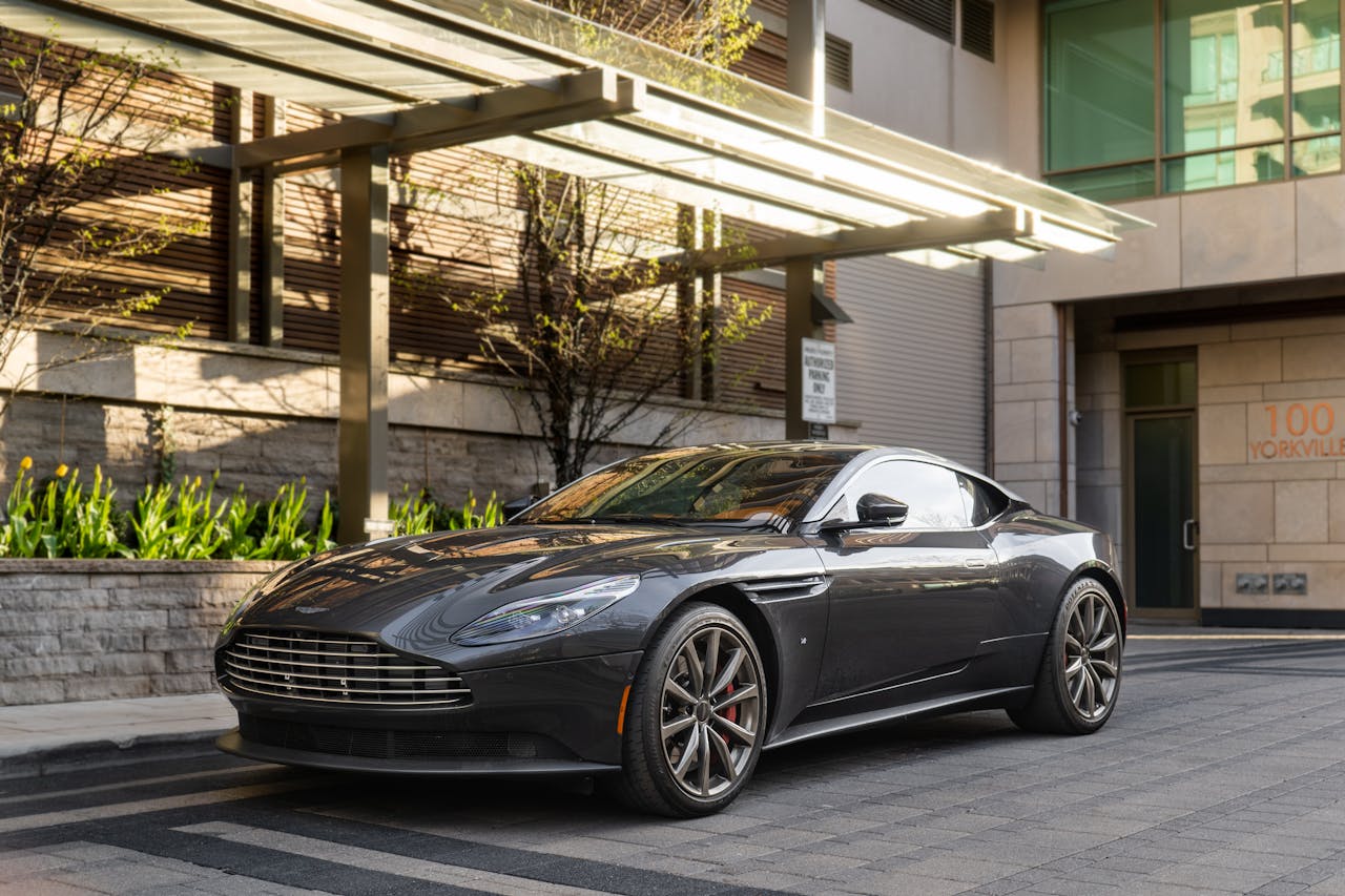 A sleek, dark grey Aston Martin DB11 parked on a paved street in front of a modern building with trees.