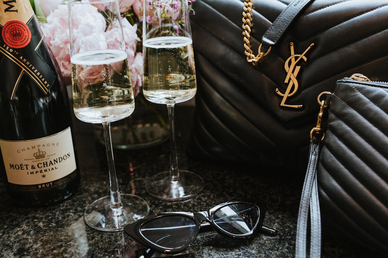 Champagne, flutes, sunglasses, and a black designer handbag on a dark surface.