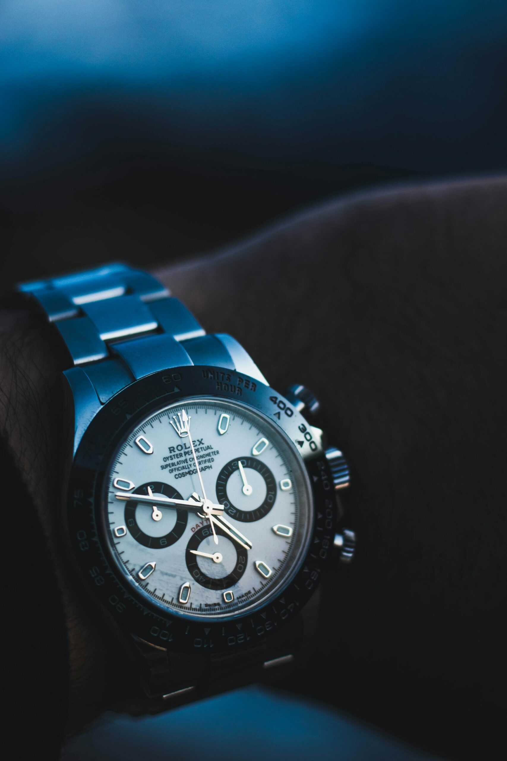 Close-up of a Rolex Daytona watch with a white dial, black subdials, and a silver metal bracelet, worn on a wrist with a dark blue background.
