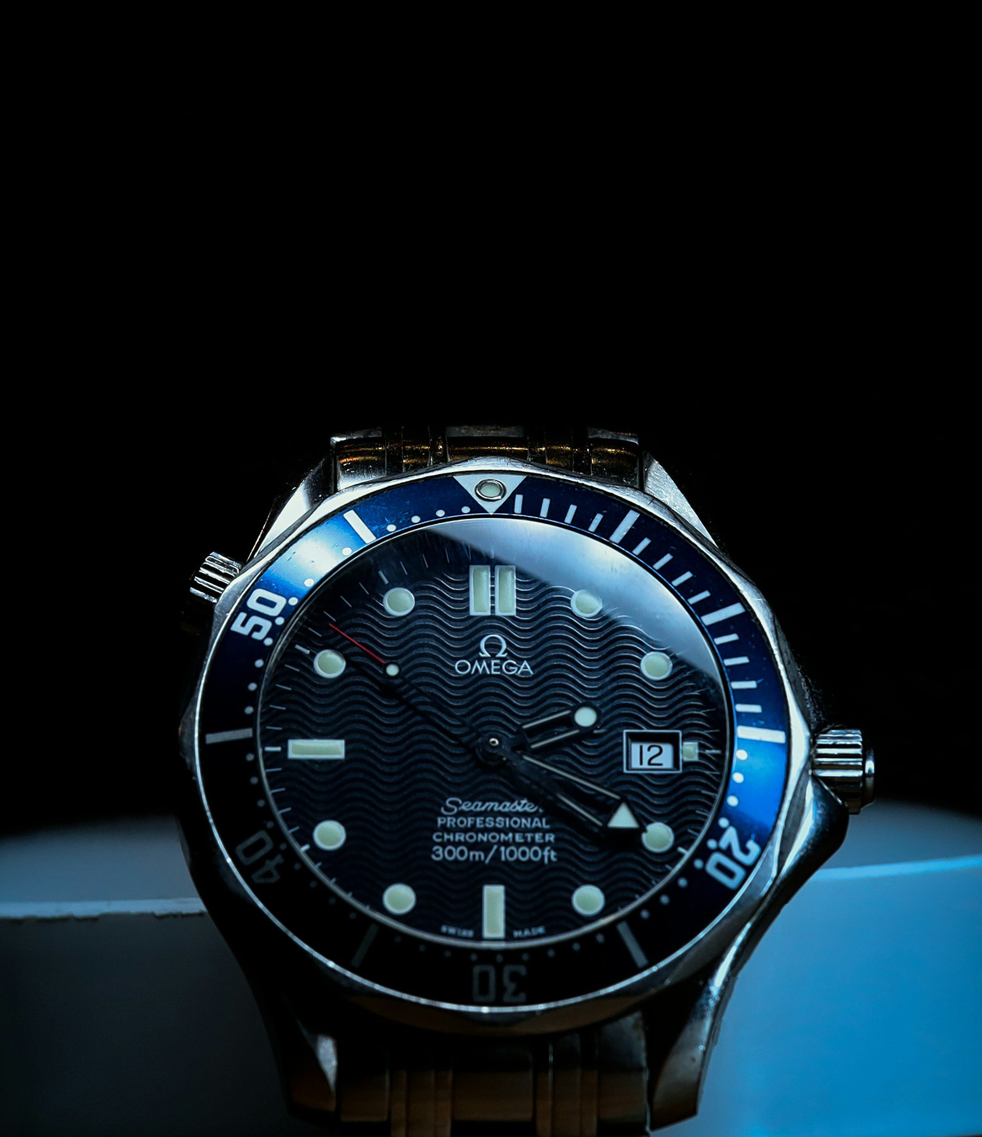 Close-up of an Omega Seamaster watch with a black dial, white hour markers, and a silver metal bracelet, displayed against a dark background.