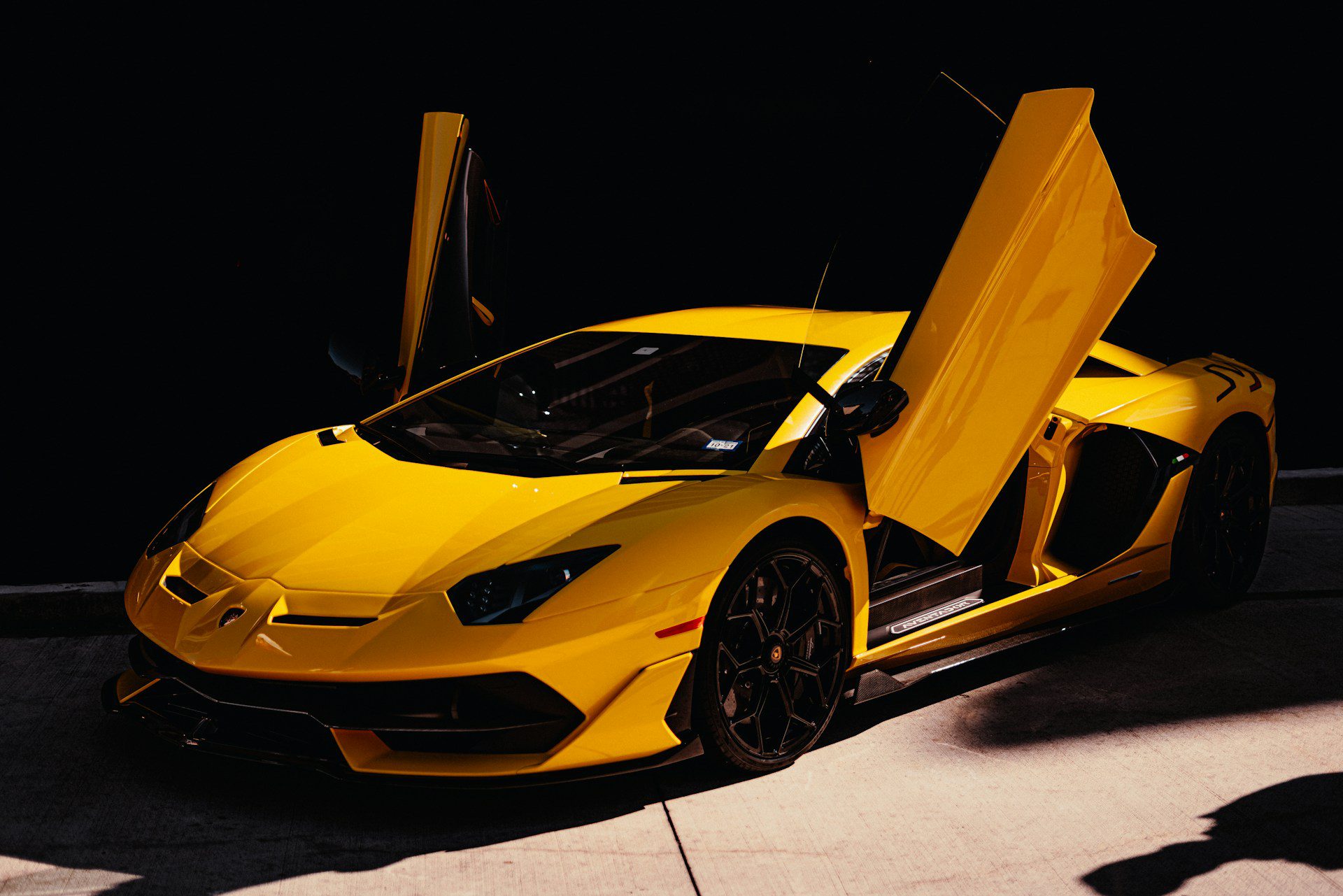 A bright yellow Lamborghini Aventador with its iconic scissor doors open, parked in a dimly lit setting.