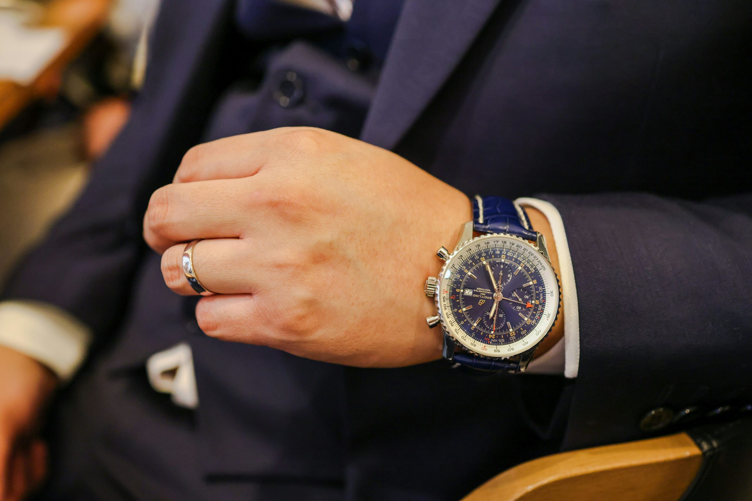 A man in a blue suit wears a blue Breitling Navitimer watch and a silver ring on his left hand.