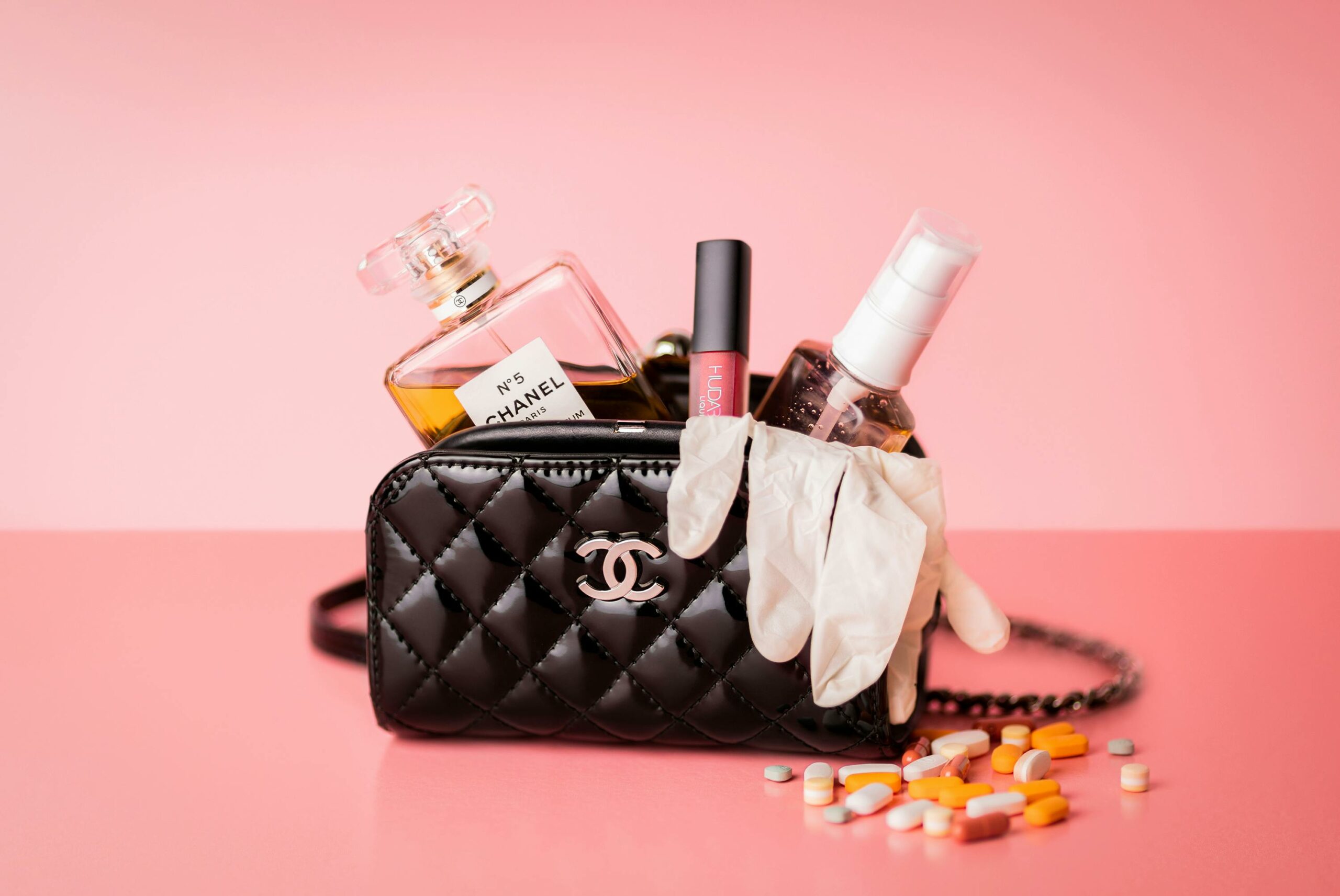 A Chanel purse with perfume, lipstick, hand sanitizer, gloves, and pills spilling out onto a pink surface.