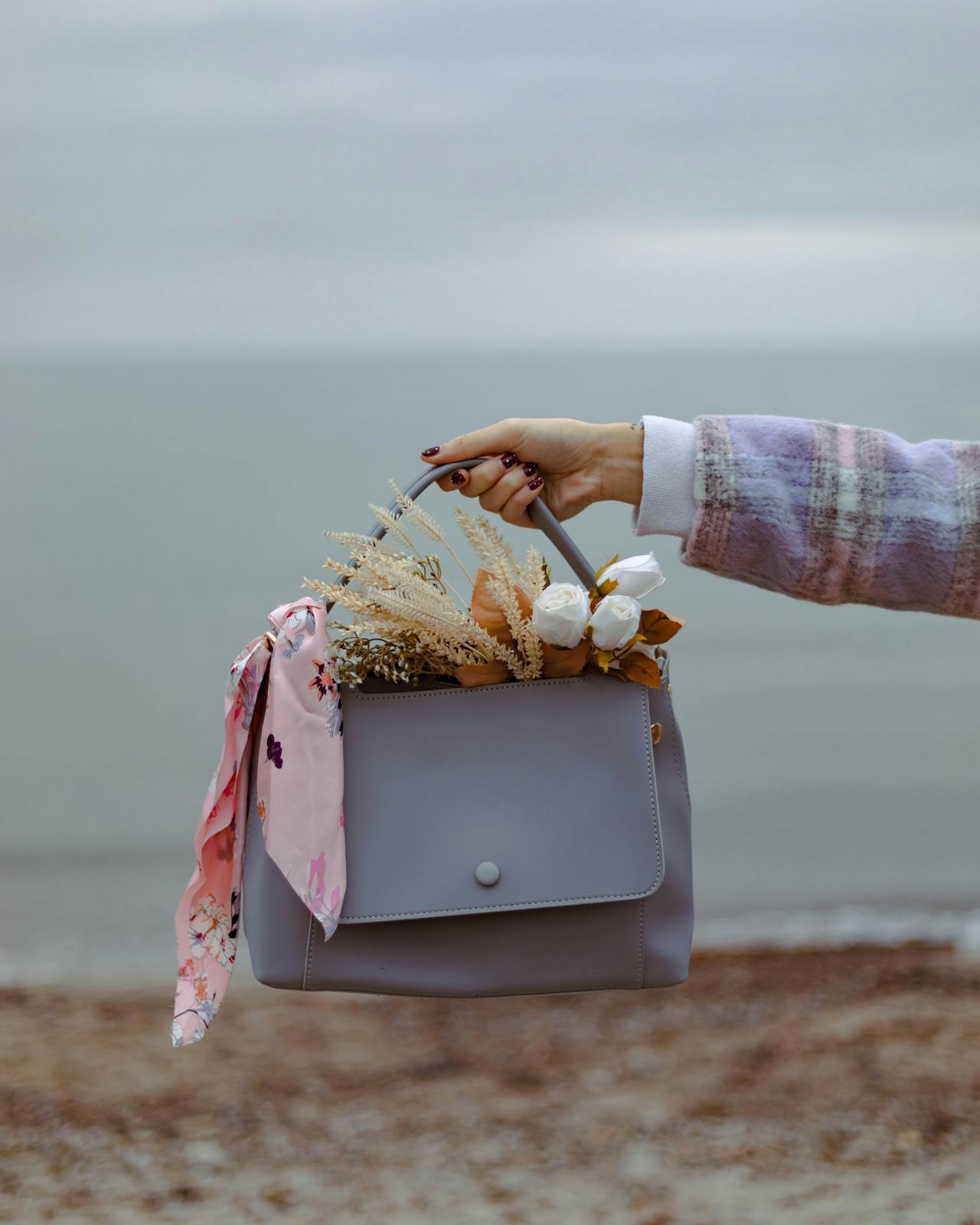 A woman holds a grey handbag filled with flowers and a pink scarf against a blurry ocean background.