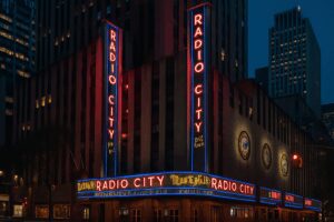 Radio City Music Hall at night with glowing red neon signs and surrounding Midtown skyscrapers