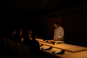 Chef preparing sushi at a dimly lit omakase counter with minimalist beige seating