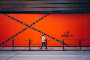 Bright orange hermes paris wall with diagonal black tape forming an x and a masked pedestrian walking along the sidewalk.