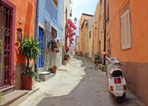 Colorful narrow street with orange, blue, and yellow houses, potted plants along the walls, and a white scooter parked on the right.