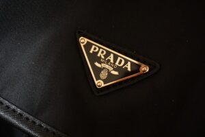 Gold-trimmed triangular prada logo patch on a black fabric surface, likely a handbag or bag exterior.