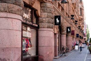 Stone-clad storefronts with massive rounded columns along a pedestrian shopping street; a bicycle is parked outside a polo ralph lauren store.