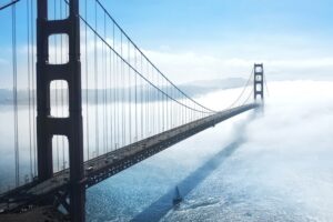 Golden Gate Bridge emerging from low-lying fog over a shimmering bay, with a sailboat beneath the span.