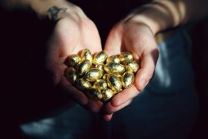 Hands cupping shiny gold foil-wrapped coins (likely chocolate coins) in dim light, center foreground.