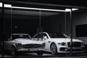 Two luxury cars in a glass-walled showroom: a vintage silver Mercedes convertible on the left and a white Bentley on the right.